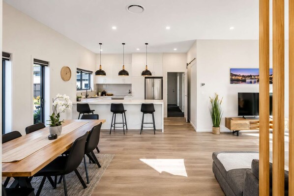 A modern kitchen with pendant lighting, a dining area with a wooden table and chairs, and a living room with a sofa and a TV.