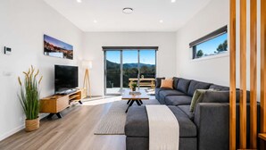 Una sala de estar moderna con un sofá grande, una televisión y vistas al exterior a través de amplios ventanales.