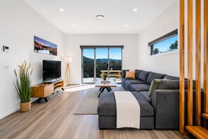 Ein modernes Wohnzimmer mit einer großen Couch, einem Fernseher und Blick ins Freie durch große Fenster.