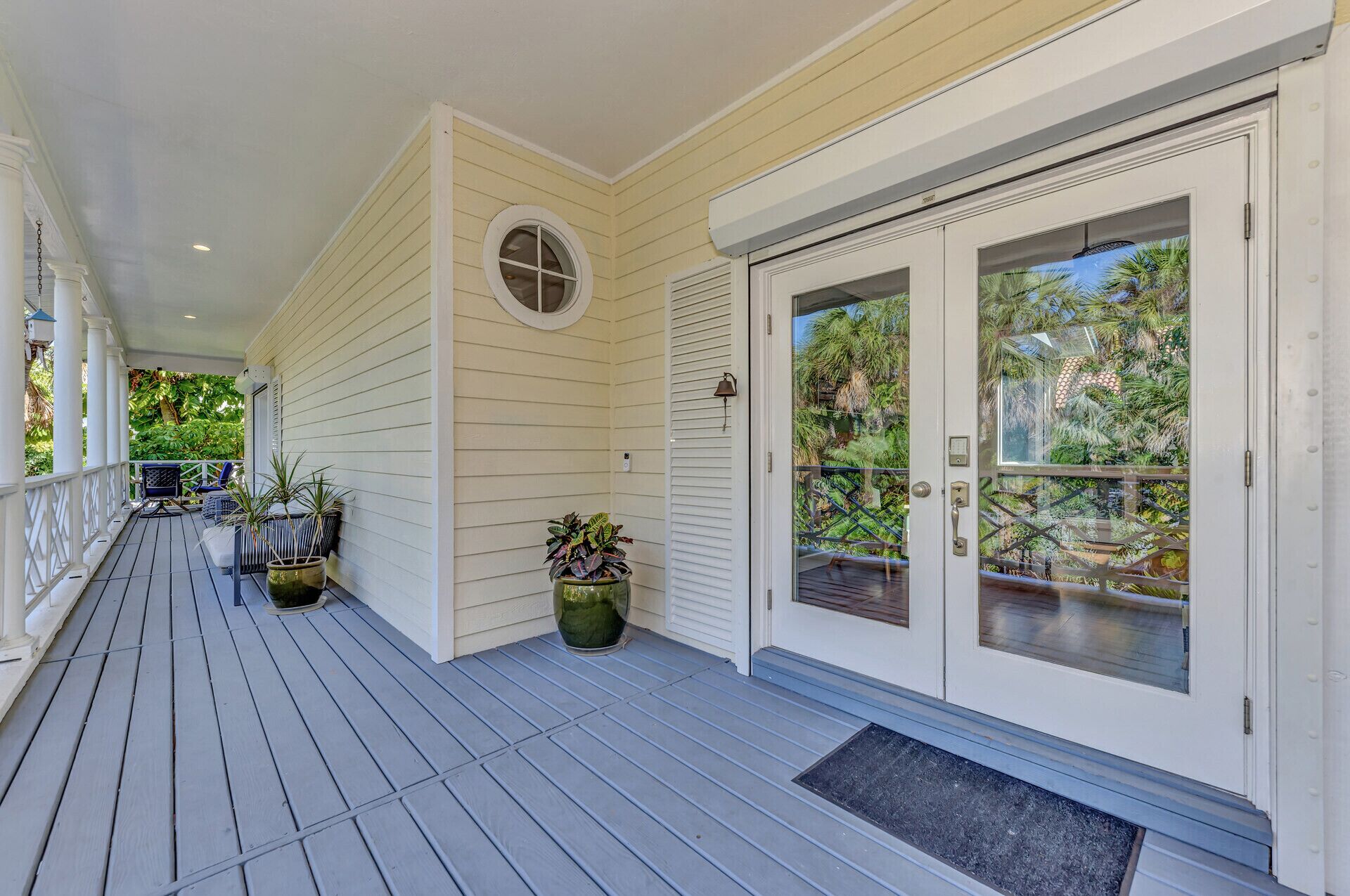 A covered porch with a wooden deck, potted plants, and a glass door leading to a balcony with a view of greenery.