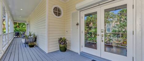 A covered porch with a wooden deck, potted plants, and a glass door leading to a balcony with a view of greenery.