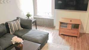 A living room with a grey sofa, a small table with a vase of flowers, a wooden cabinet, and a large TV on the wall. - Mathilda Street Trio - City View - 2 Queen Beds (Pittsburgh)