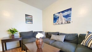 A living room with a dark brown coffee table, a grey sofa, a small side table with a potted plant, and a framed picture of a building on the wall. - Mathilda Street Trio - City View - 2 Queen Beds (Pittsburgh)