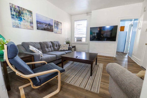A modern living room with a grey sofa, a wooden coffee table, and a flat-screen TV mounted on the wall. - Mathilda Street Trio - Stylish 2BR Apt (Pittsburgh)