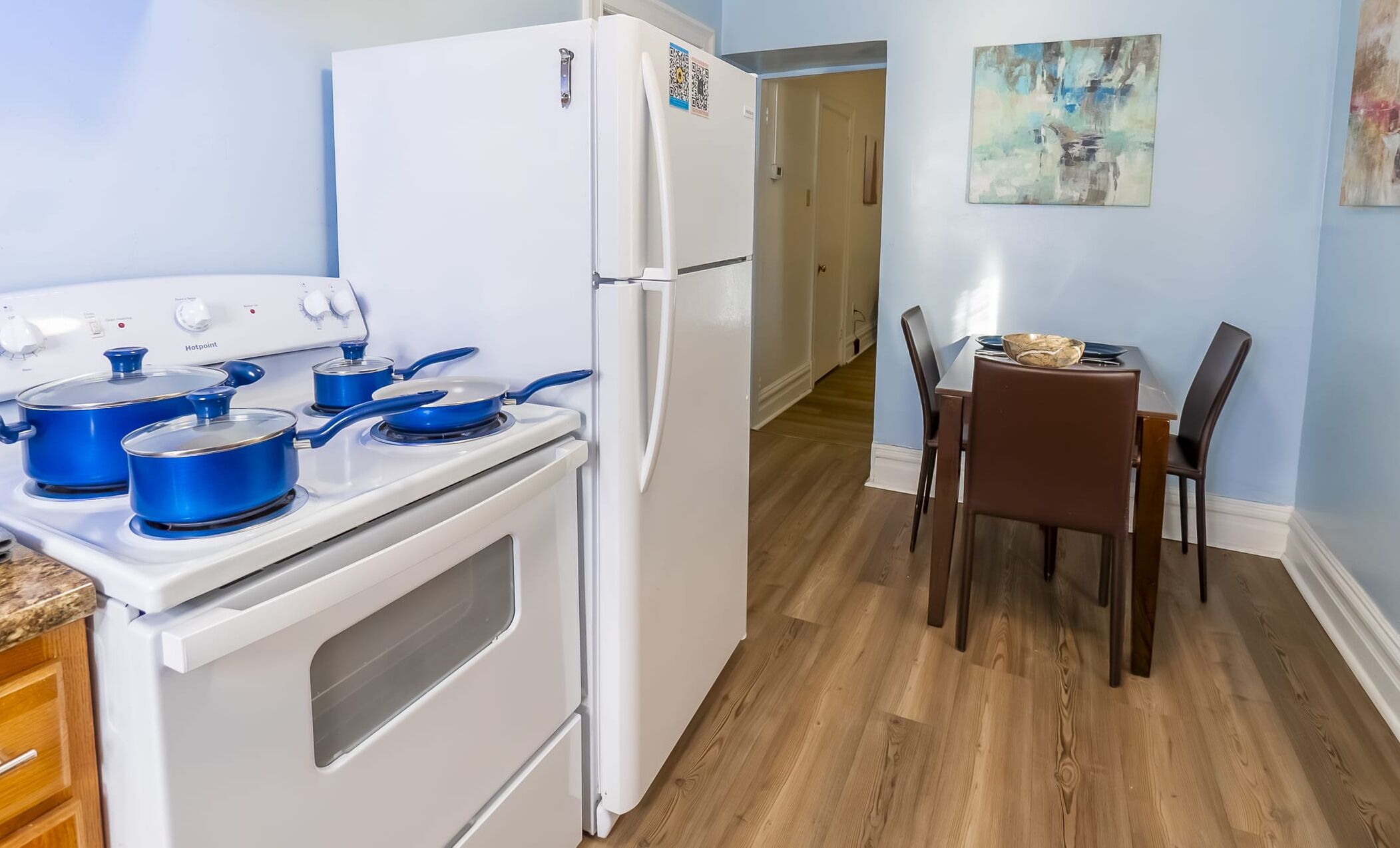 A kitchen with a white refrigerator, a stove with blue pots, a dining table with chairs, and a painting on the wall.