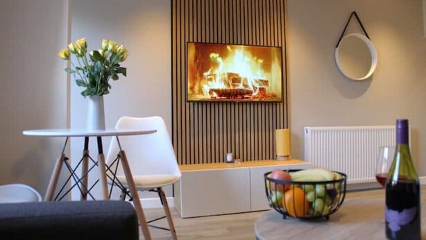 A modern living room with a TV mounted on a wooden panel, a white table with a fruit bowl, a white chair, and a vase with yellow flowers. - Modern Stylish Apartment with central access (Perth)