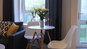 A modern living room with a dark-colored sofa, a white chair, a small round table with a vase of yellow flowers, and a potted plant on a windowsill. - Modern Stylish Apartment with central access (Perth)