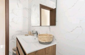 Un baño con pared de mármol, tocador de madera, lavamanos, espejo y cesta de mimbre.