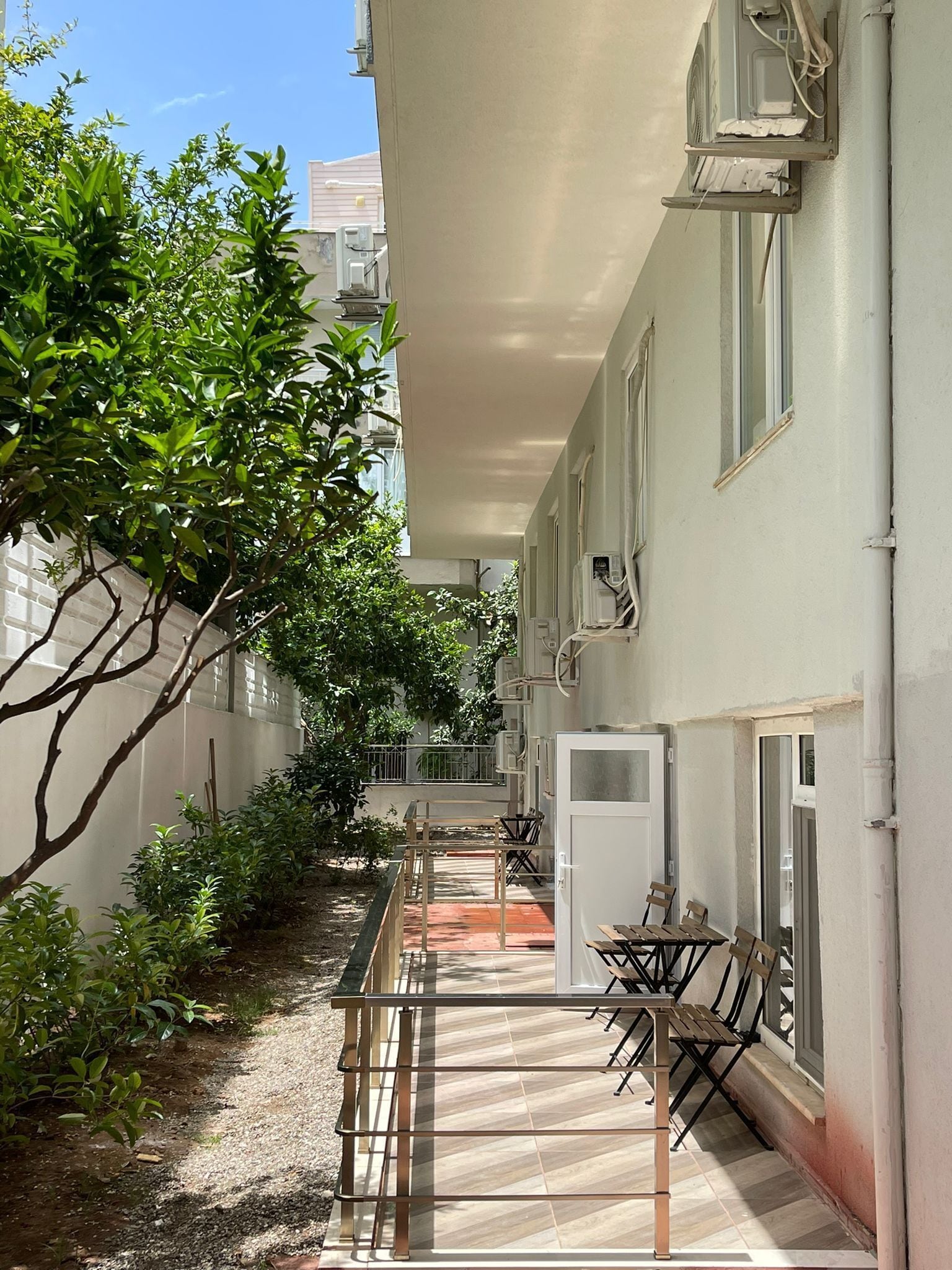 A narrow outdoor corridor with a metal railing, a small table with chairs, and a door.