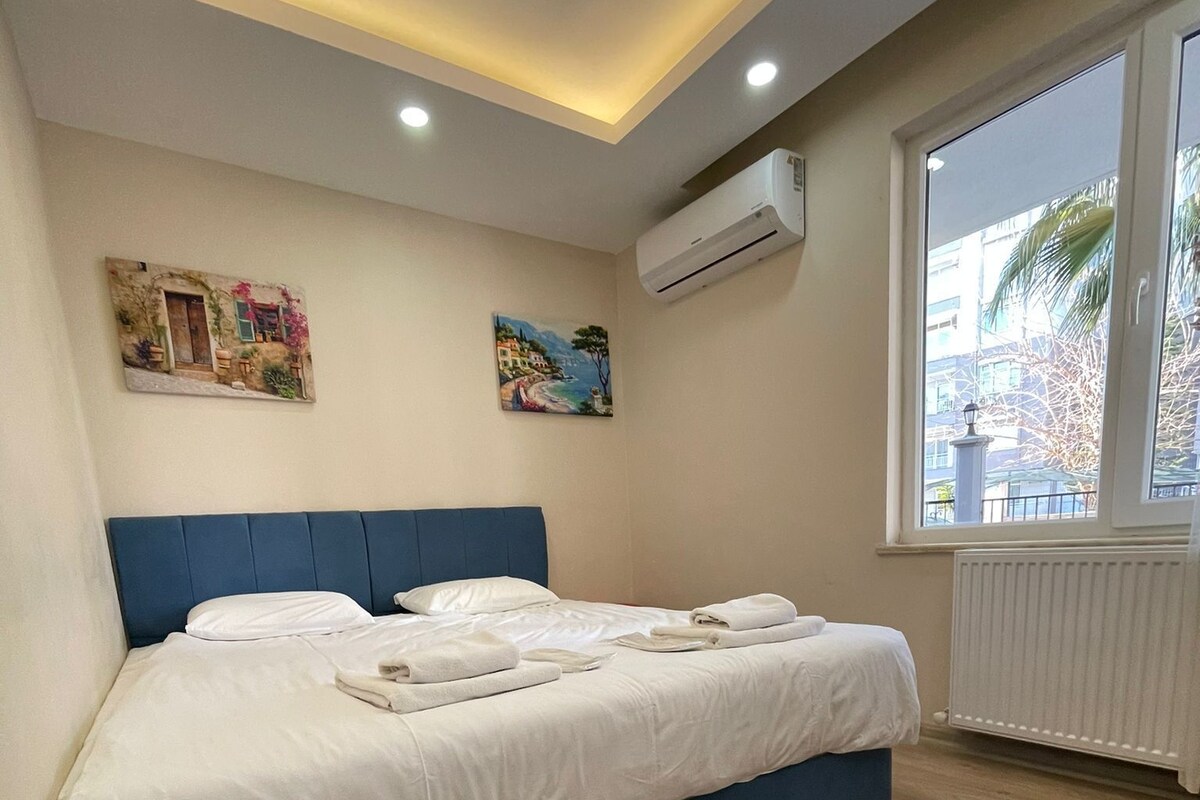 Un dormitorio con una cama grande, una lámpara en el techo, un equipo de aire acondicionado y una ventana con vistas a edificios y palmeras.