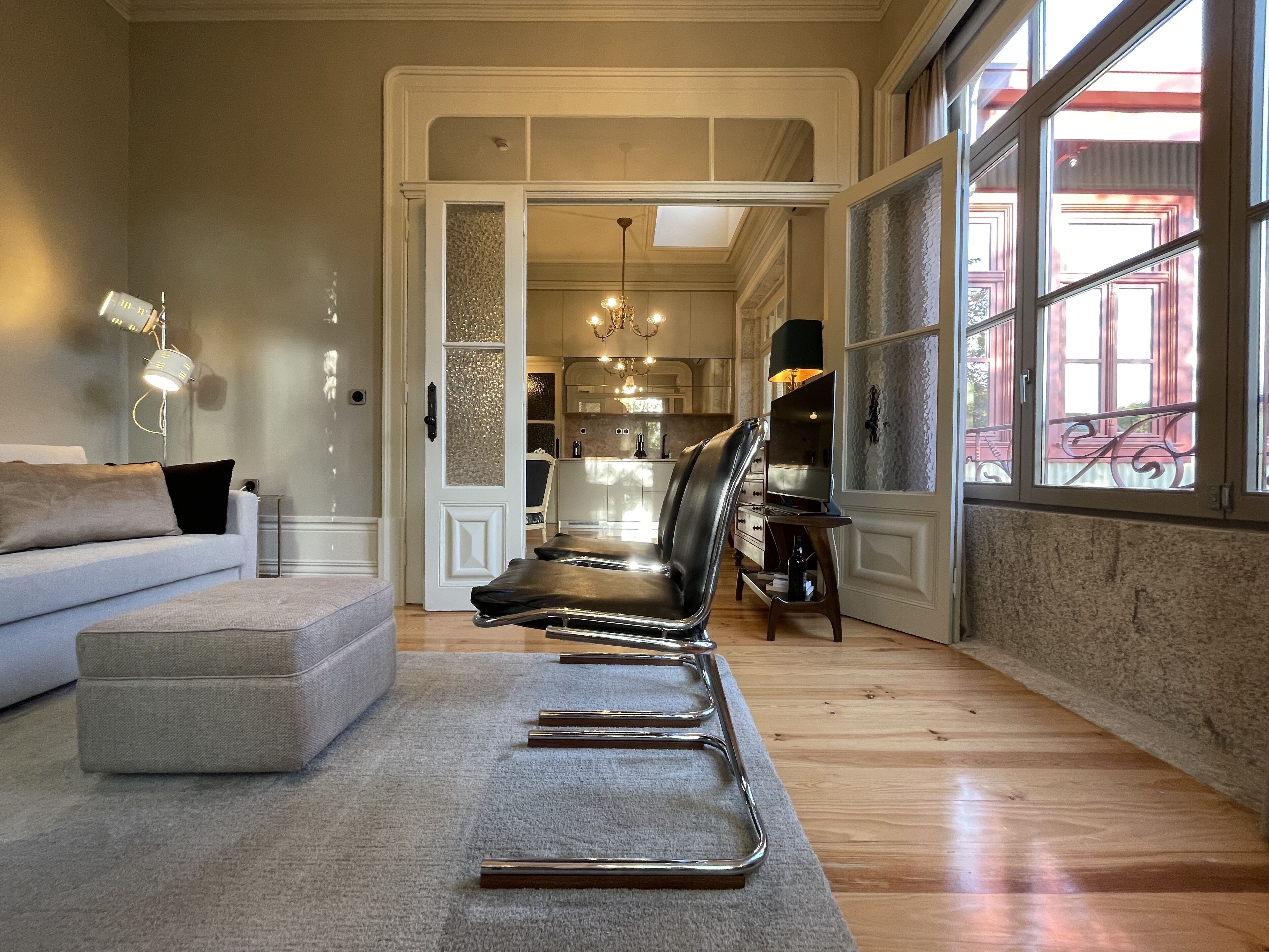 Un salon moderne avec un canapé gris, un pouf gris et un fauteuil en cuir noir. Le sol est en bois, il y a un tapis gris et une grande fenêtre donnant sur un immeuble rouge.