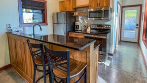 Una cocina con armarios de madera, un refrigerador de acero inoxidable, un microondas y una estufa. Hay una zona de bar con taburetes y una ventana con persianas.