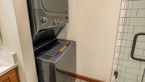 Un lavadero pequeño con lavadora y secadora de carga frontal, una pequeña encimera y una zona de ducha con azulejos.