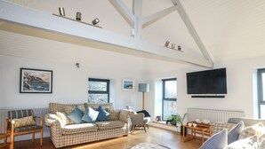 A spacious living room with a high ceiling, a large window, a sofa set, a television, and a radiator. - Plas Elyn (Beaumaris)