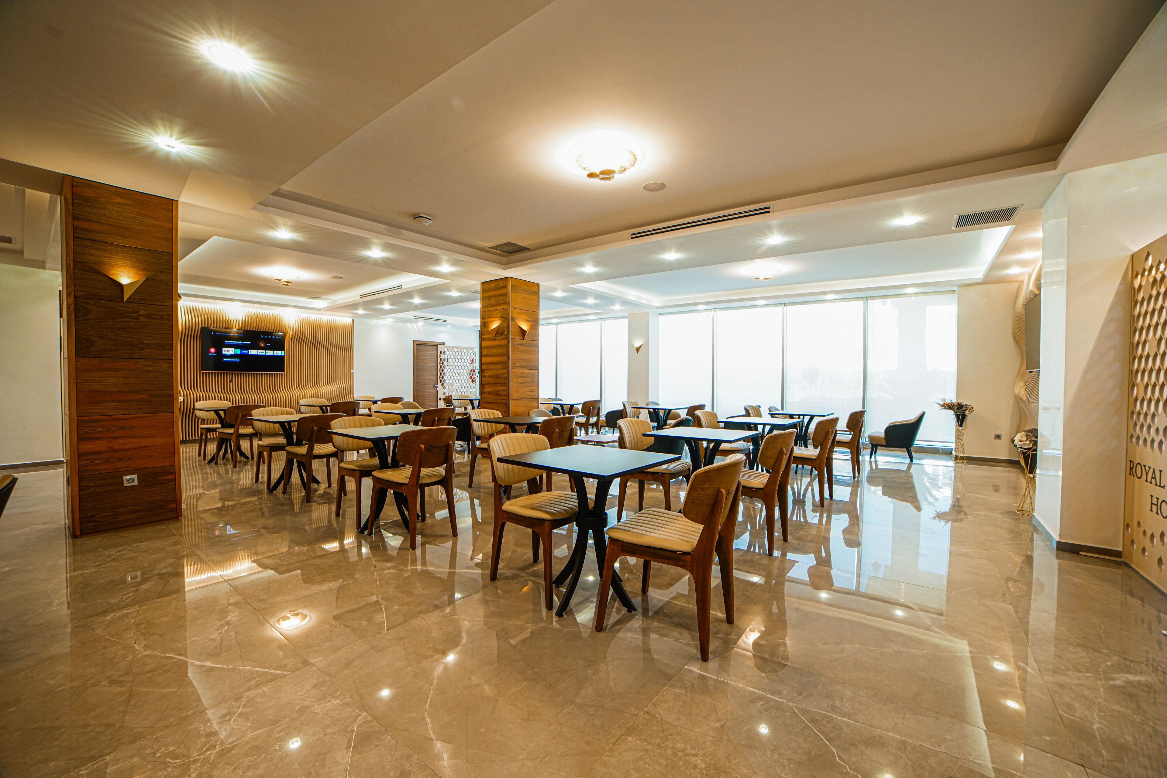 A spacious dining area with multiple tables and chairs, a flat-screen TV mounted on the wall, and large windows allowing natural light.