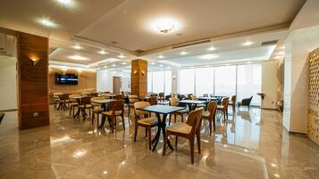 A spacious dining area with multiple tables and chairs, a flat-screen TV mounted on the wall, and large windows allowing natural light.