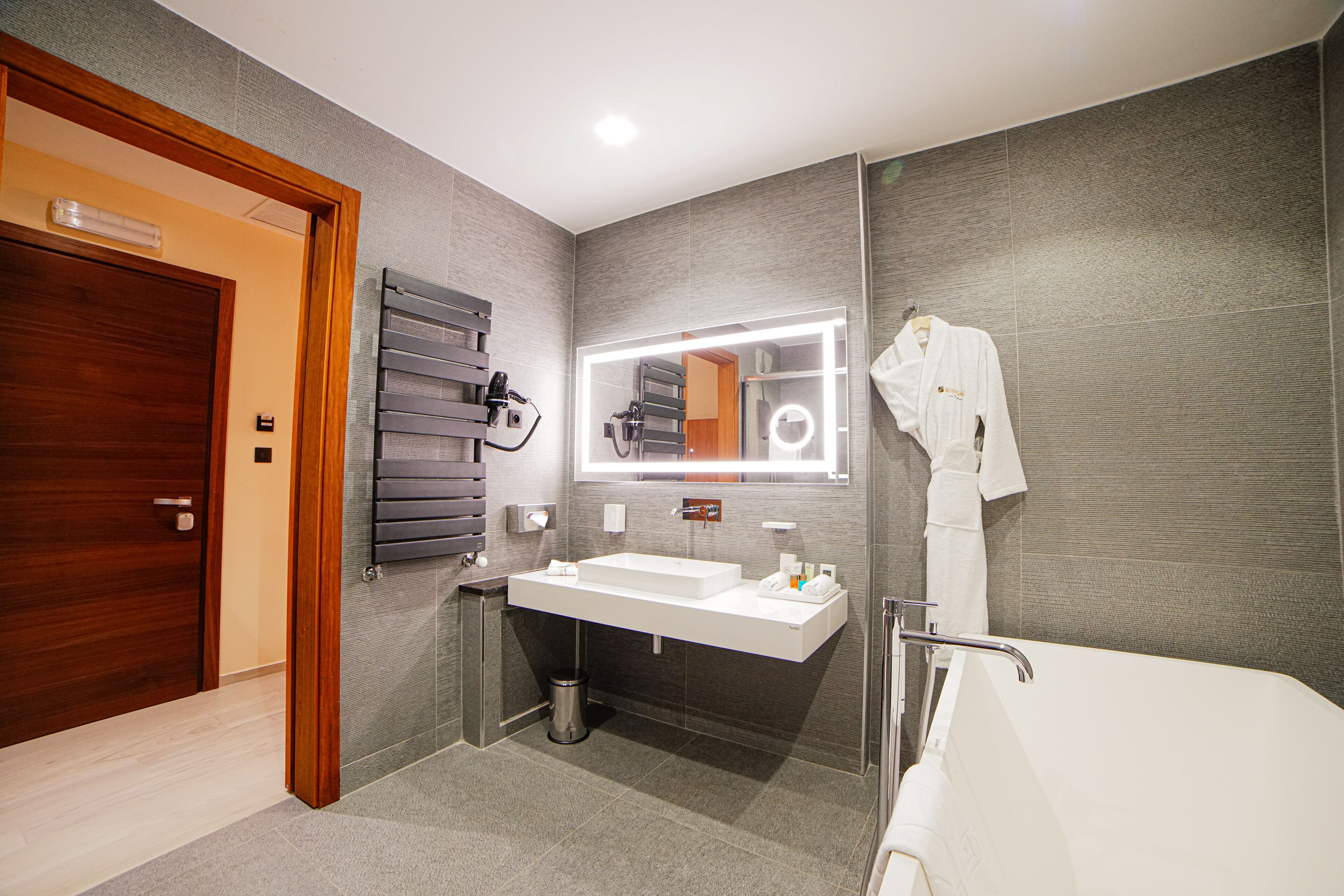 Un baño moderno con puerta de madera, un espejo grande con iluminación, un lavabo blanco y una bañera.