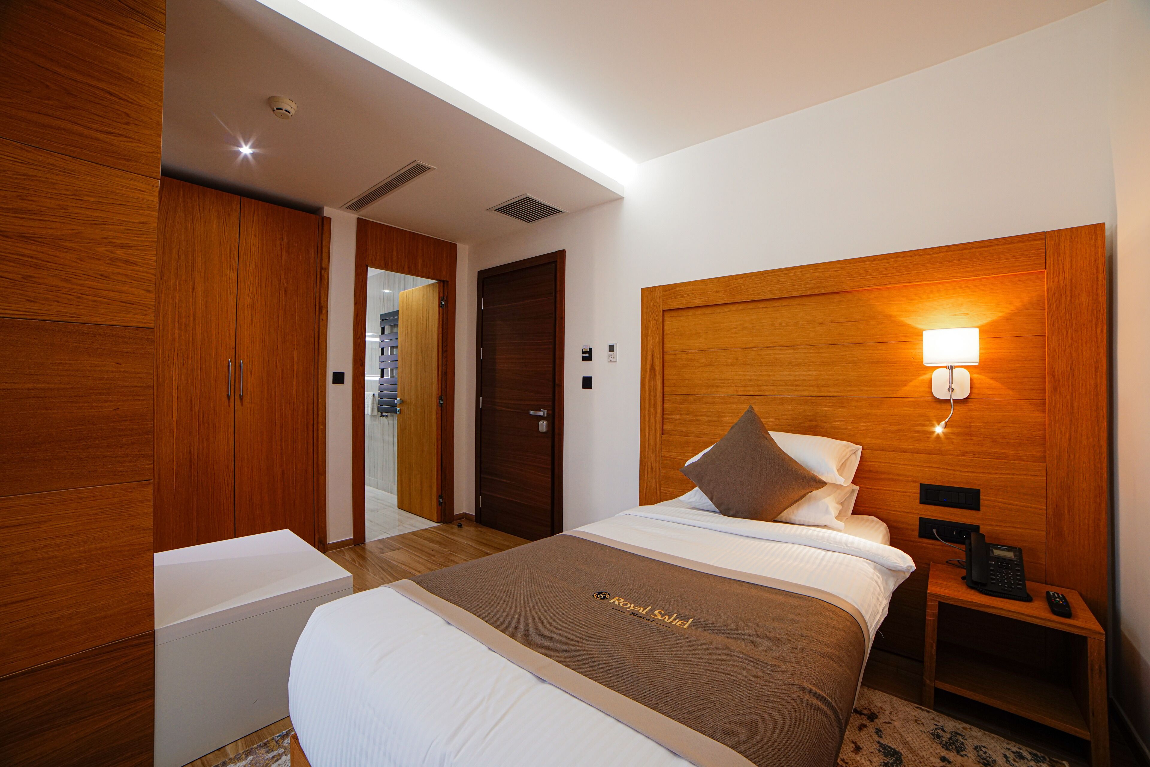 Habitación de hotel con una cama grande, armario de madera, mesita de noche con lámpara y una pequeña mesa con teléfono.
