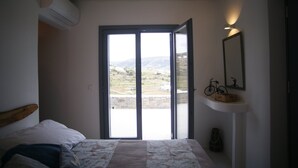 A bedroom with a bed, a window with a view of a landscape, a wall-mounted shelf with a bicycle, and an air conditioning unit. - Krotiri View of Paros (Paros)
