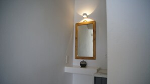 A bathroom with a mirror, a light fixture above it, and a small shelf with a decorative item. - Krotiri View of  Paros (Paros)