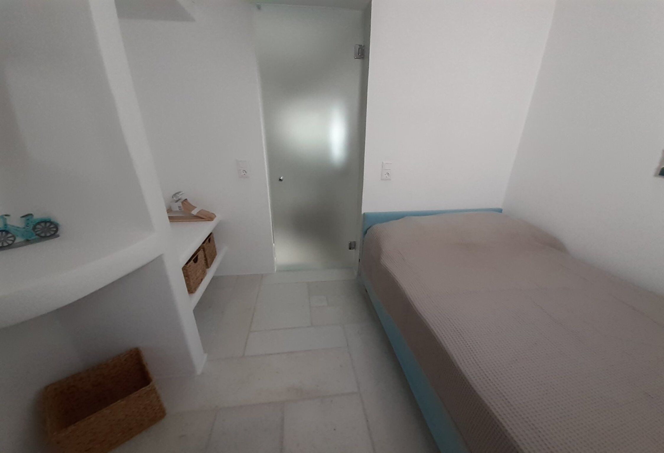 Ein schmales Zimmer mit einem Bett, einem Schuhregal und einer Tür zu einem weiteren Raum.