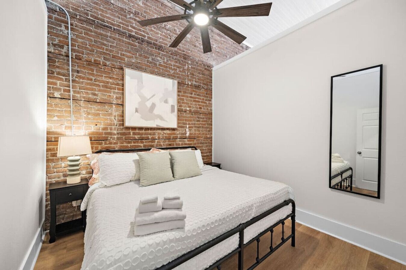 Ein Schlafzimmer mit Backsteinwand, Deckenventilator, Bett mit weißer Bettwäsche, Nachttisch mit Lampe und einem großen Spiegel an der Wand.