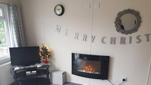 A cozy living room with a fireplace, a television on a stand, a small decorated Christmas tree, and a grey rug. - Lovely 2-bed Chalet at Robinhood Retreat Free Park (Newark)