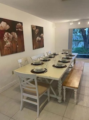 Un comedor con una larga mesa dispuesta para seis personas, sillas blancas y dos cuadros en la pared.