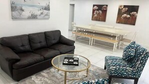 Ein Wohnzimmer mit einer schwarzen Ledersofa, einem gemusterten Sessel, einem Glastisch und einem Marmorboden. An der Wand hängt ein Gemälde mit Vögeln und ein weiteres mit Blumen.