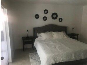 Un dormitorio con una cama, dos mesitas de noche, una mesita auxiliar, un reloj en la pared y una puerta.