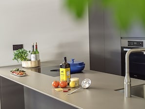Una cocina moderna con encimera de acero inoxidable, horno integrado y un lavabo. Hay dos botellas de vino, una planta y algunos alimentos en la encimera.