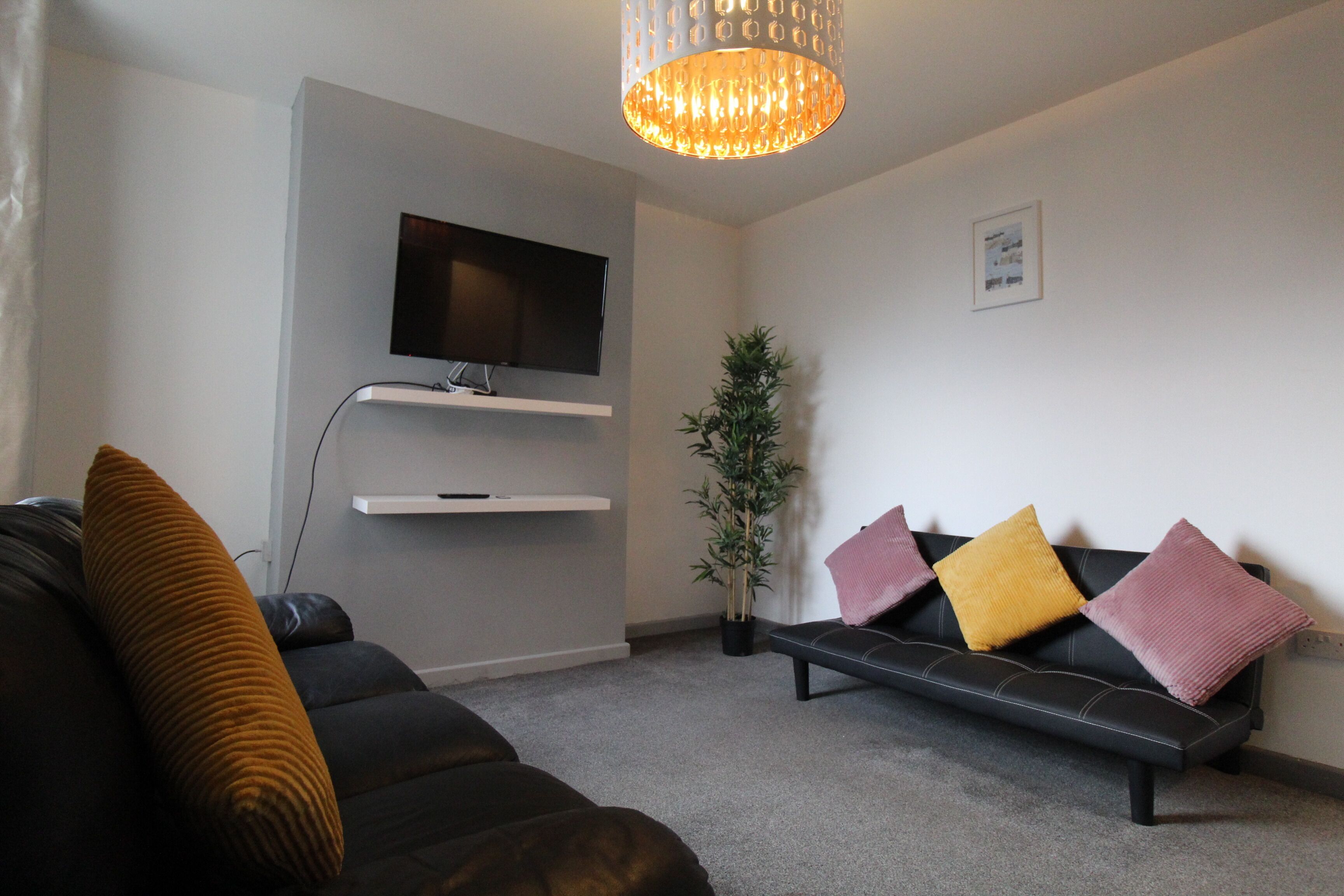 A modern living room with a black sofa, a bench, a flat-screen TV mounted on the wall, a potted plant, and a decorative light fixture.