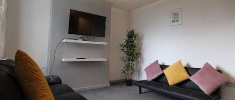 A modern living room with a black sofa, a bench, a flat-screen TV mounted on the wall, a potted plant, and a decorative light fixture.