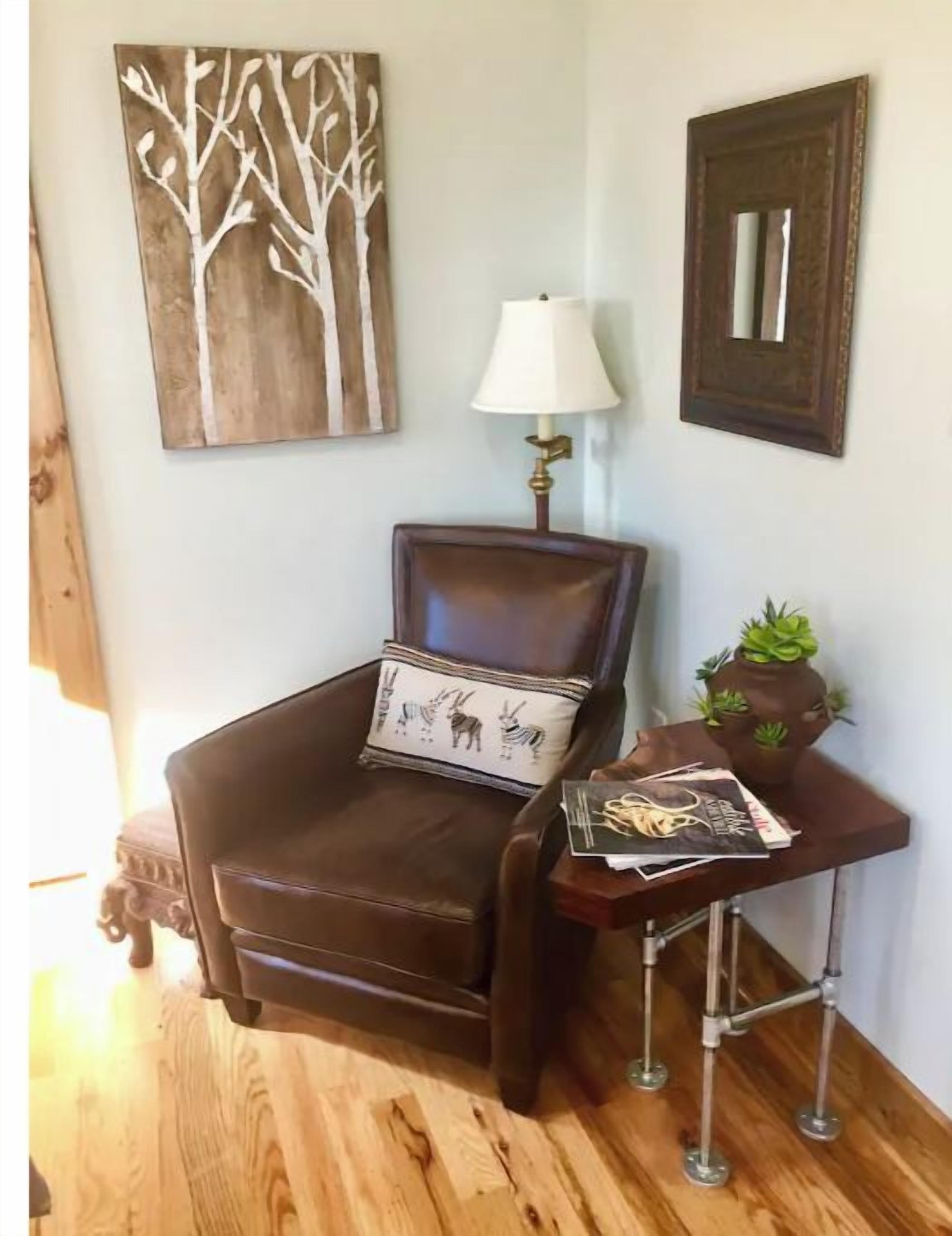 A corner room with a brown leather chair, a small table with a lamp, a framed mirror, and a wall art piece.