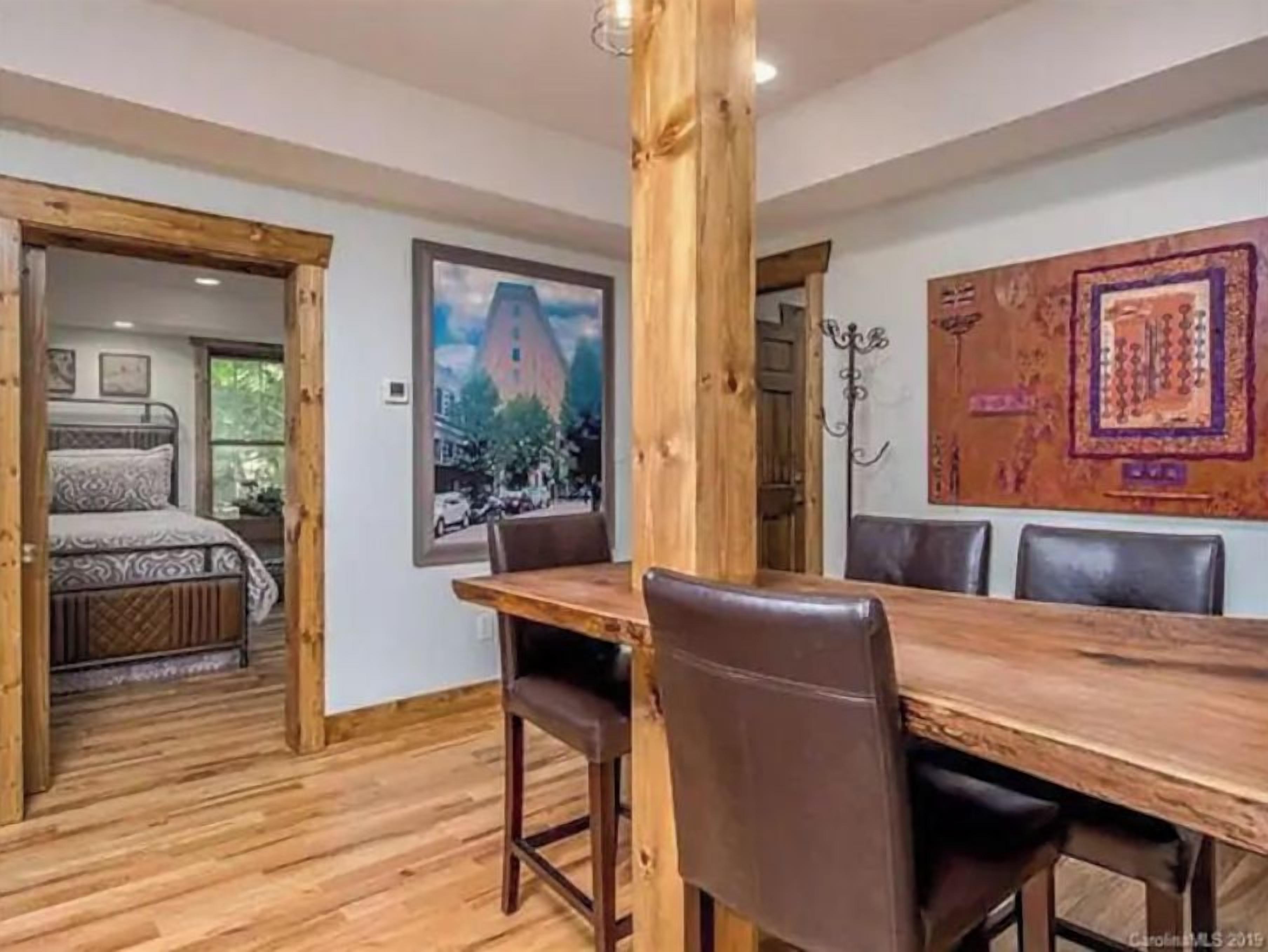 A room with a wooden dining table and chairs, a bed with patterned bedding, and a large framed picture on the wall.