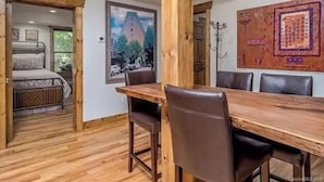 A room with a wooden dining table and chairs, a bed with patterned bedding, and a large framed picture on the wall.