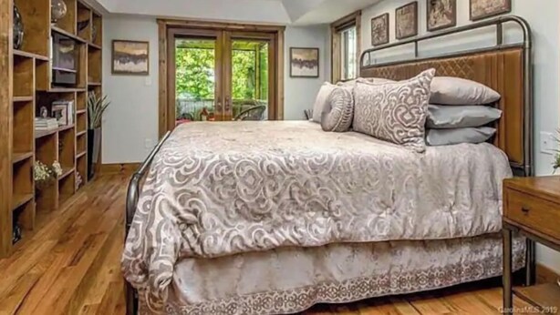 A bedroom with a large bed, wooden floor, built-in bookshelf, and a window with a view of greenery.