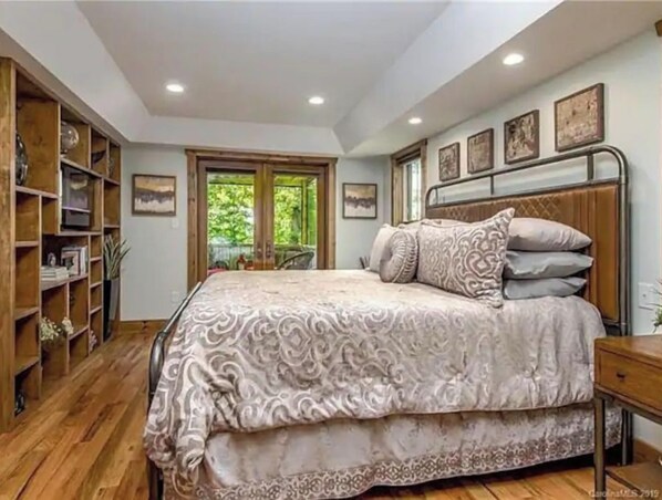 A bedroom with a large bed, wooden floor, built-in bookshelf, and a window with a view of greenery. - Near Biltmore Village · Fireplace · Treetop Porch (Asheville)