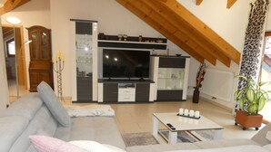 Een moderne woonkamer met een grote bank, een salontafel en een televisie. Er is een houten plafond en een kast met glazen deuren.