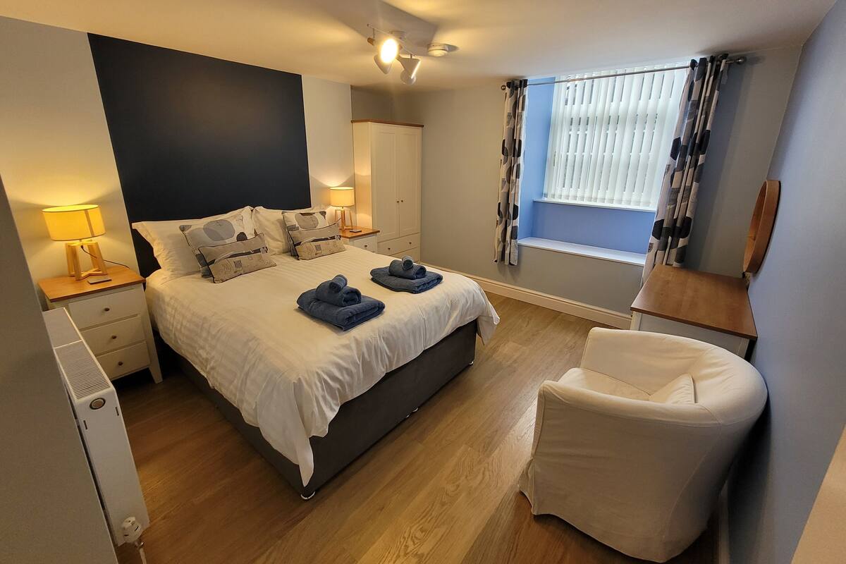 Un dormitorio ordenado con cama, mesitas de noche, una silla y una ventana con cortinas.