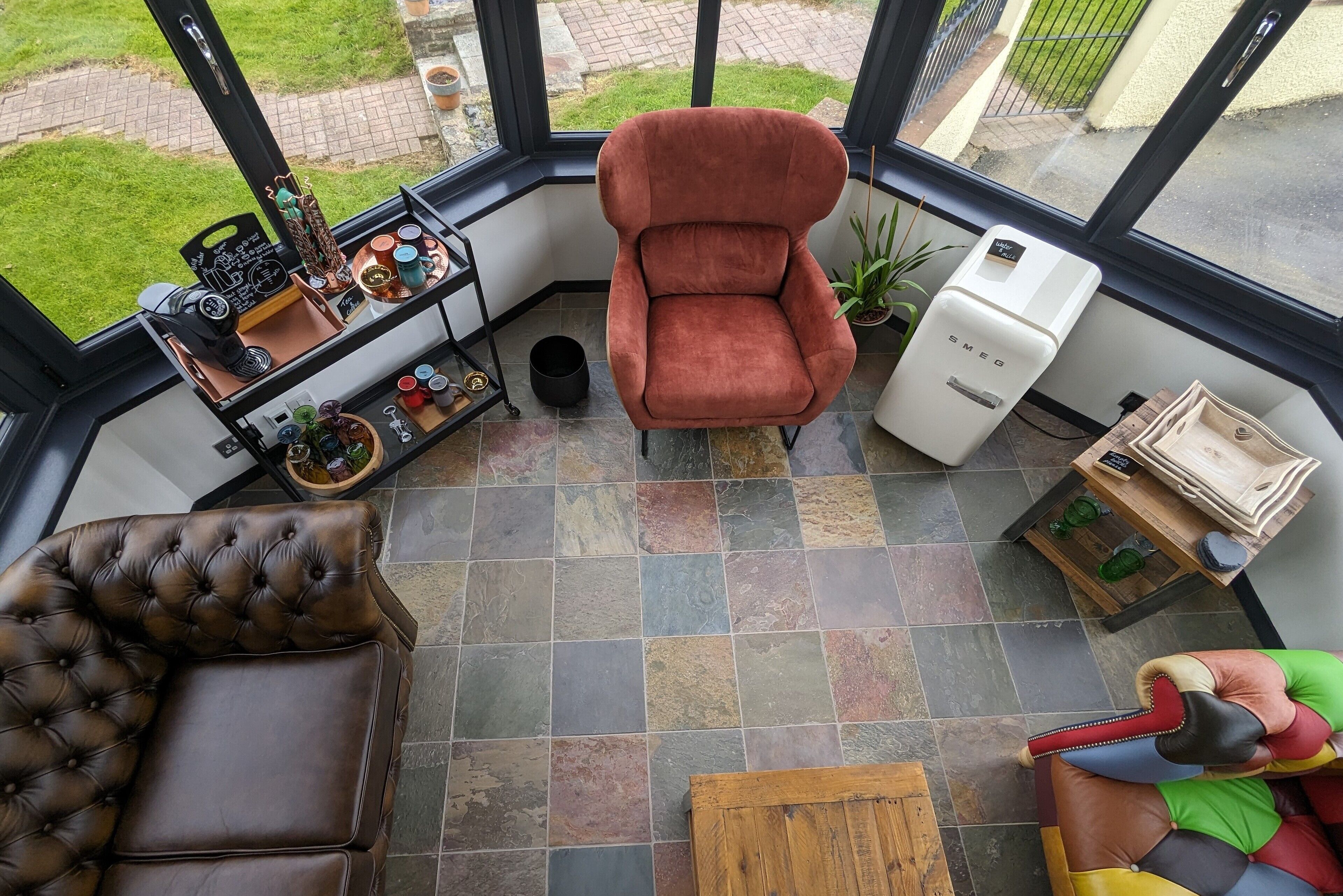 Una habitación acogedora con un sofá de cuero marrón, un sillón de terciopelo rojo y una silla tapizada y colorida. Se ve un electrodoméstico blanco, y la habitación tiene ventanas grandes con vistas a un jardín.
