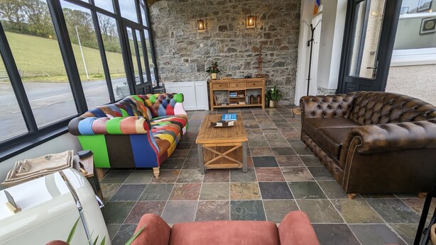 A spacious room with a stone wall, large windows, a colorful sofa, a brown leather couch, and a wooden coffee table. - Llys Aeron Guest House (Aberaeron)