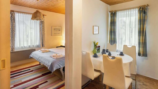 Une pièce comprenant un lit, une table à manger avec des chaises et une fenêtre avec des rideaux.