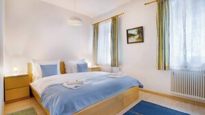 A hotel room with a bed, bedside tables, a lamp, a window with curtains, a radiator, and a painting on the wall.