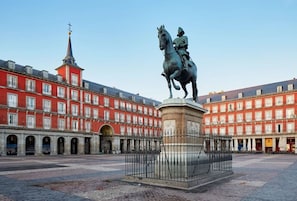 Une place où se trouve une statue d’un homme à cheval, entourée de bâtiments rouges dotés de nombreuses fenêtres, et où l’on aperçoit, au loin, le clocher d’une église.