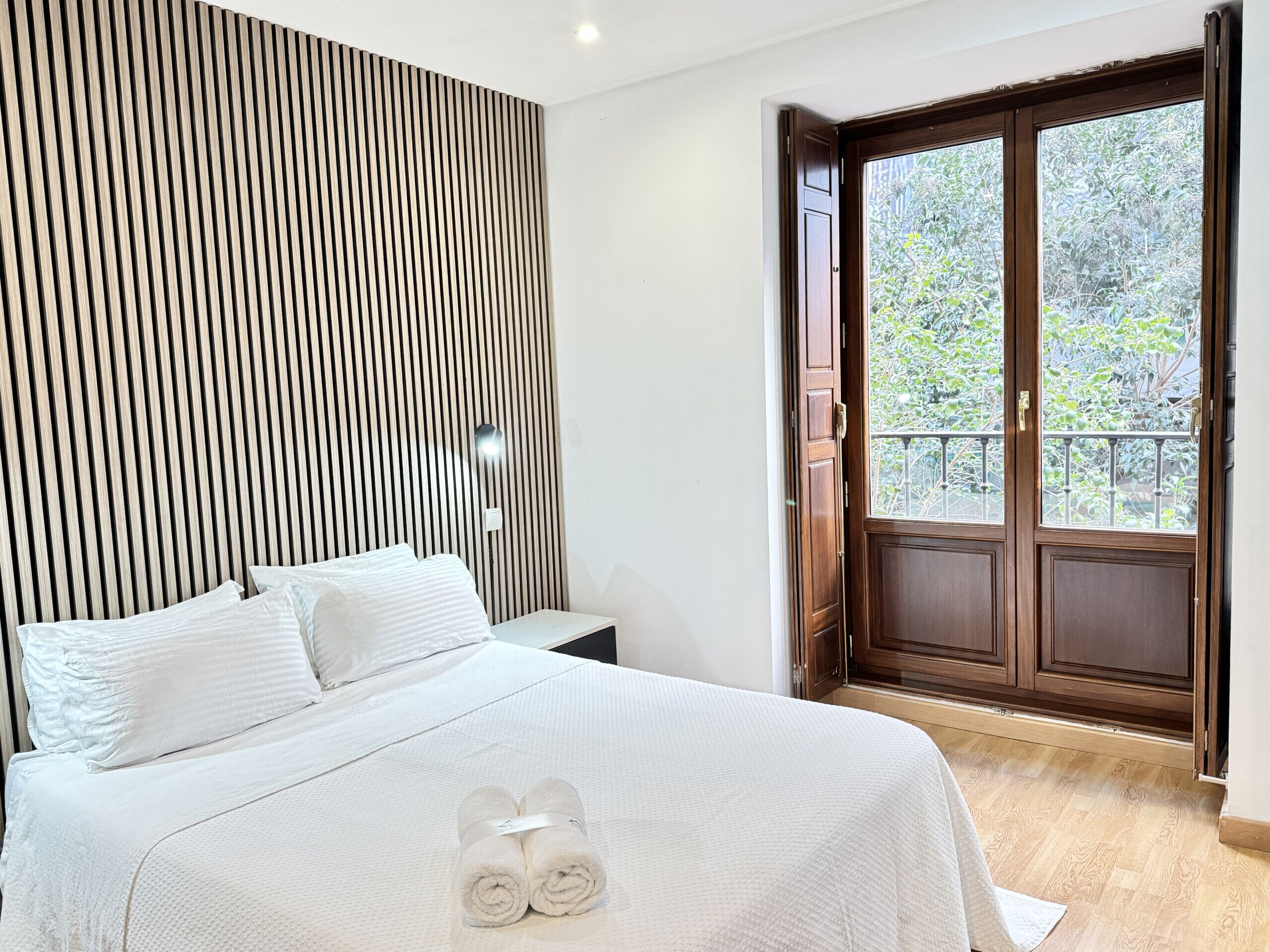 Ein Schlafzimmer mit einem Bett, einem Holz-Schlafkopf, einem Nachttisch und einem Fenster mit Blick auf Grünflächen.