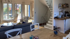 Amplia sala de estar con una mesa de comedor de madera, asientos cómodos, una escalera que lleva a una cocina y ventanas grandes que ofrecen vistas al exterior.