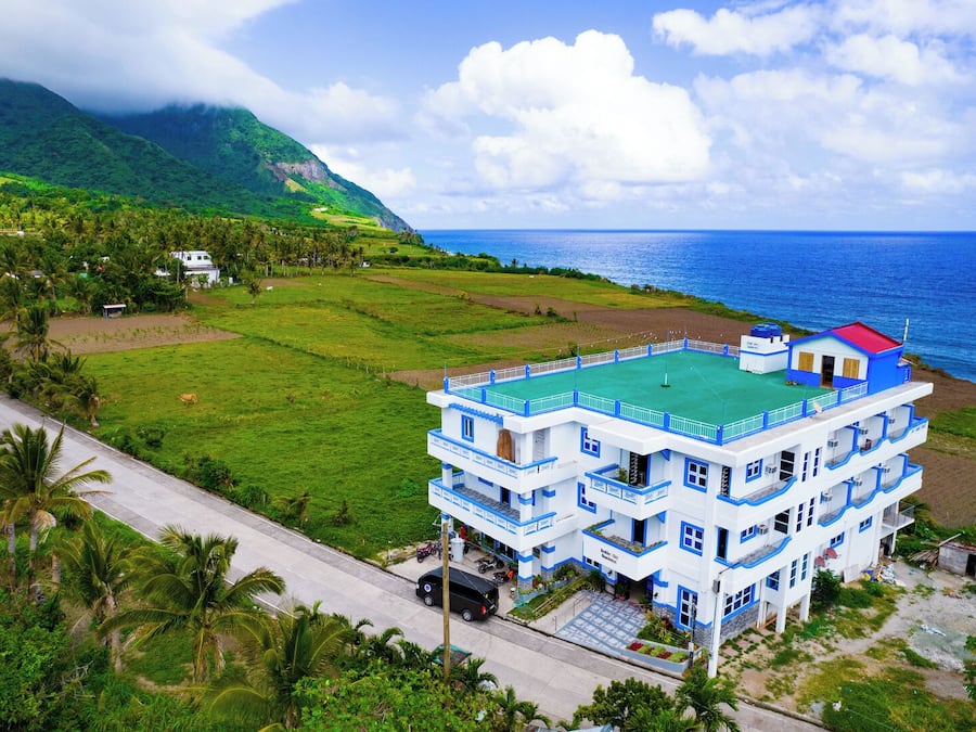 Un hotel de varios pisos con una terraza en la azotea de color verde, situado cerca de la costa y con vistas al océano y montañas.
