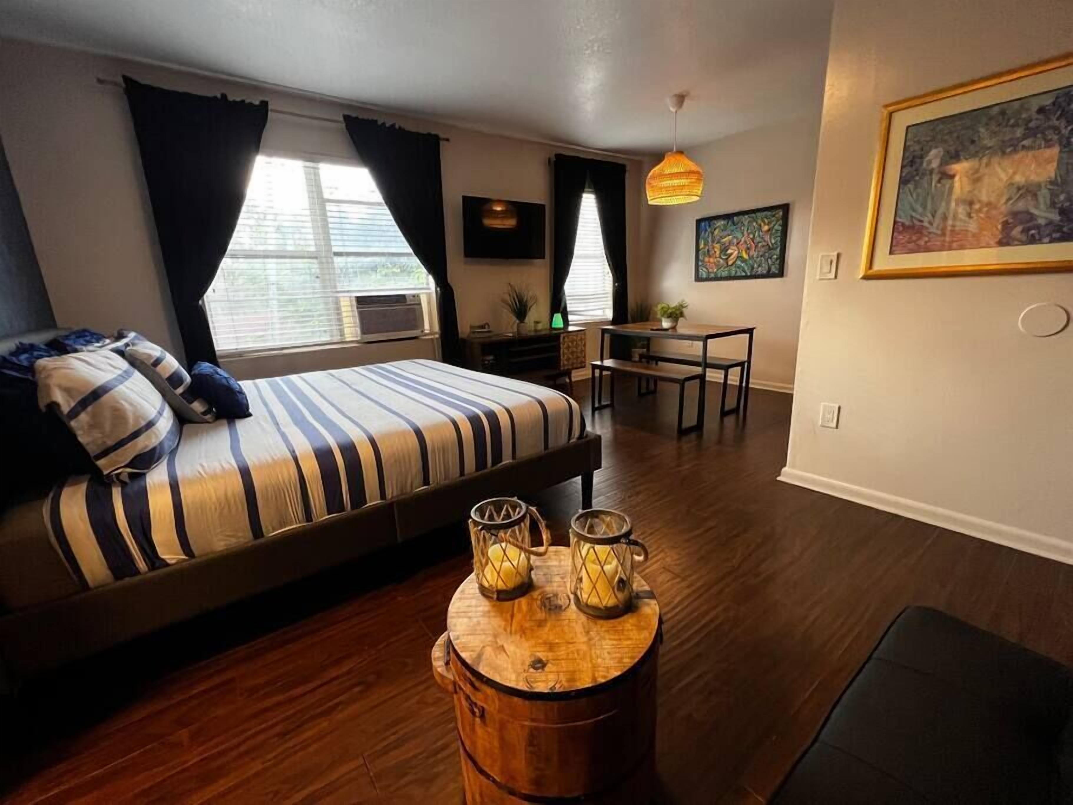 A bedroom with a bed, a desk, and a dining table. There are windows with curtains, a hanging light fixture, and a painting on the wall.