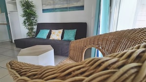 Een moderne woonkamer met een donkere bank, een witte salontafel en een rieten fauteuil. Er staat een grote plant en er hangt een schilderij van een strand aan de muur.
